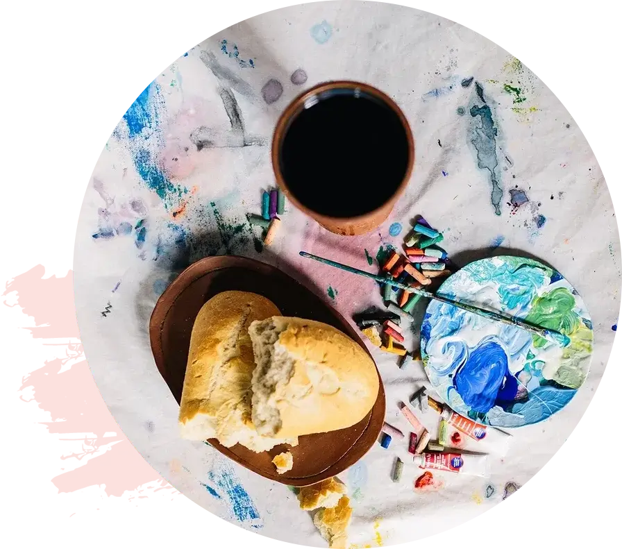 A table with bread on a plate, a cup of dark liquid, and artist supplies including paints and crayons scattered on a white surface, embodying the theme of healing through art.